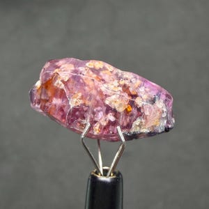 May include: A raw, translucent purple gemstone with internal crystalline inclusions and orange-toned flecks, held securely by a silver-toned metal pronged display stand. The stone features a jagged, irregular surface texture with visible fractures.