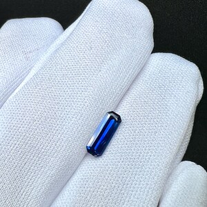 May include: A deep blue gemstone with a rectangular shape, cut into a baguette style. The gemstone is displayed on a white fabric background.