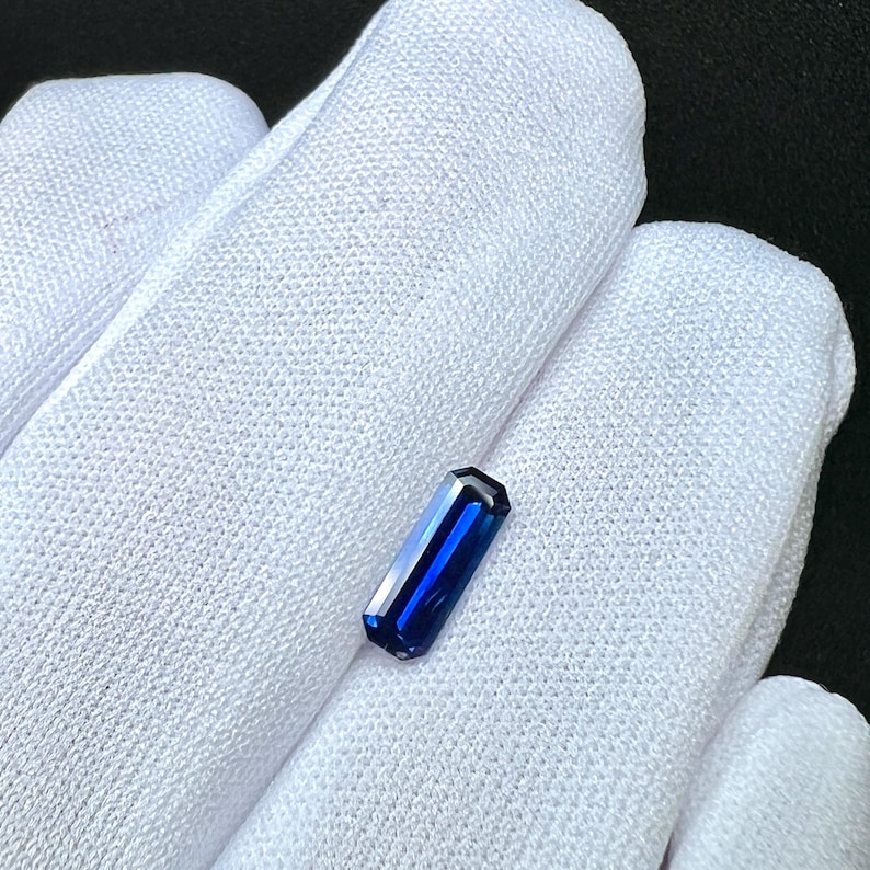 May include: A deep blue gemstone with a rectangular shape, cut into a baguette style. The gemstone is displayed on a white fabric background.