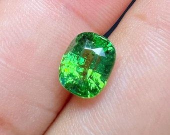 May include: A vibrant, faceted green gemstone with a cushion cut. The gem is a bright, vivid green color and appears to be suspended by a black string. The gemstone is held in a person's hand.