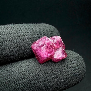 Könnte beinhalten: Ein Cluster aus rohen, ungeschliffenen rosa Edelsteinen. Die Kristalle haben eine raue, strukturierte Oberfläche und eine lebendige, gesättigte Farbe. Die Edelsteine werden vor einem dunklen, strukturierten Hintergrund gehalten.