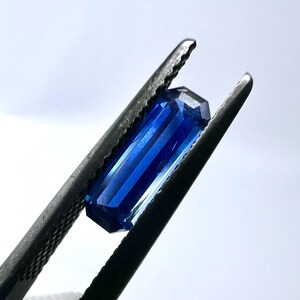 May include: A deep blue gemstone with a rectangular cut, held in tweezers.