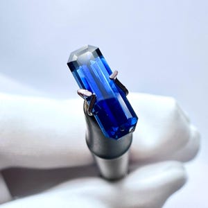 May include: A deep blue gemstone with a rectangular cut, held in a metal clamp.