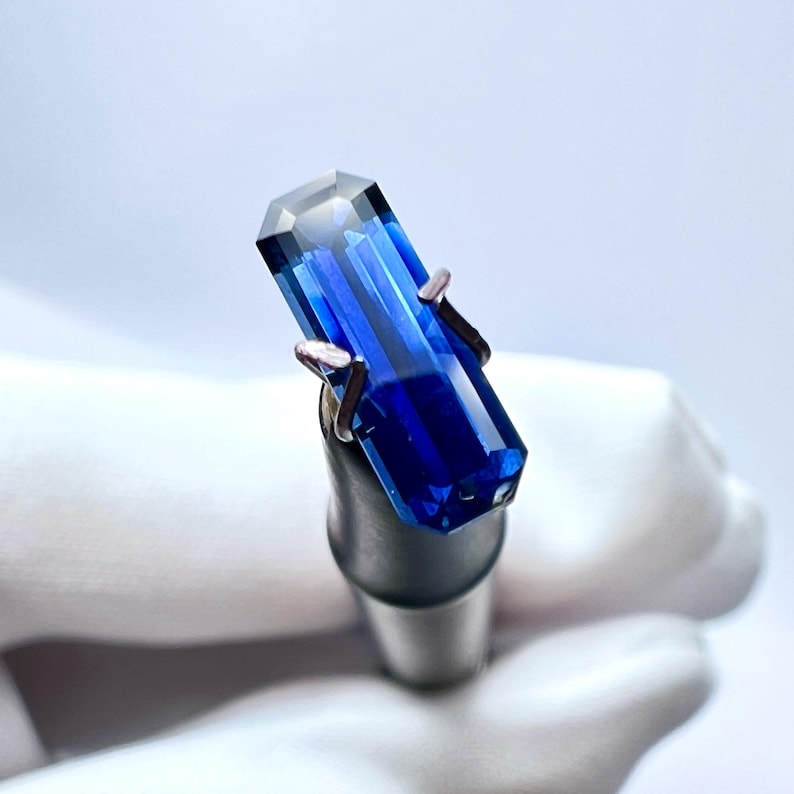 May include: A deep blue gemstone with a rectangular cut, held in a metal clamp.
