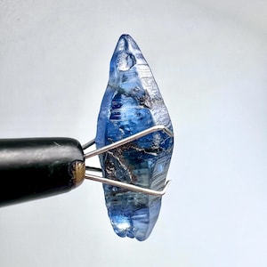 Puede incluir: Un cristal de zafiro azul en bruto sujeto con pinzas metálicas. El zafiro tiene una punta afilada y una superficie rugosa y texturizada. El color varía de azul claro a azul oscuro, con algunas zonas casi negras. El cristal se encuentra sobre un fondo blanco liso.