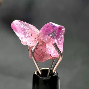 以下が含まれることがあります： 金属製のツールで保持された、生のピンク色の宝石のクローズアップ。 宝石は、ピンクと紫のさまざまな色合いを持つ結晶構造をしています。 ツールは黒いベースと銀色の金属製の爪を持っています。