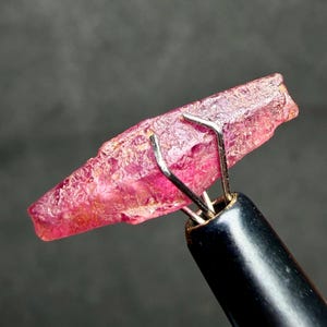 May include: A raw, elongated pinkish-red gemstone crystal with a textured, uneven surface held securely by a metal prong tool. The crystal displays translucent qualities and natural facets, showcasing a vibrant, deep magenta hue throughout its structure.