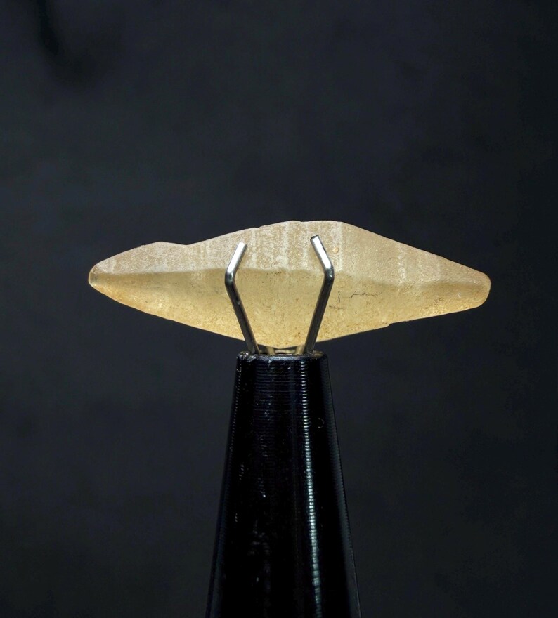 Puede incluir: Una gema transl&uacute;cida de color amarillo claro est&aacute; sujeta por una pinza de metal plateado sobre un soporte negro. La gema tiene una forma rugosa e irregular con un extremo ligeramente puntiagudo. El fondo es negro liso.