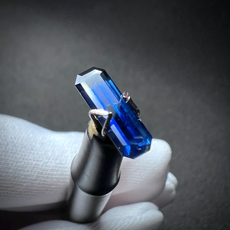 May include: A deep blue gemstone with a rectangular cut, held in a metal setting. The gemstone is being examined under a magnifying glass.