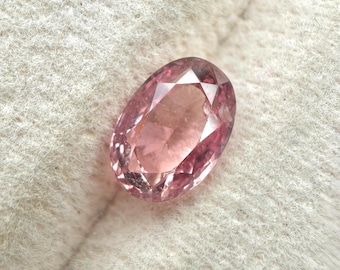 May include: A faceted, oval-shaped gemstone with a soft pink hue. The gem displays internal facets that catch and reflect light, creating a sparkling effect. The stone is set against a neutral, textured background.