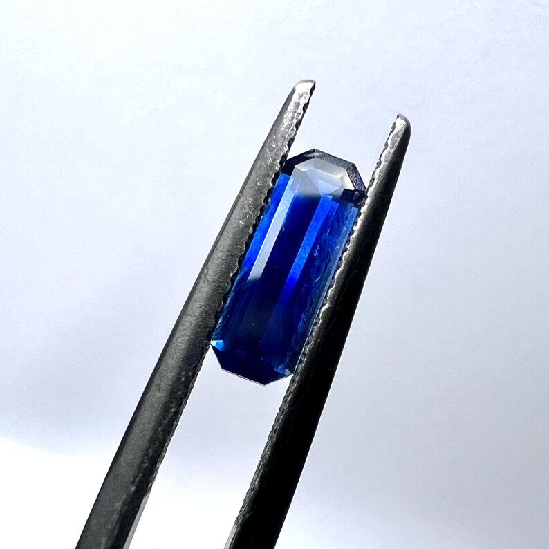 May include: A deep blue gemstone with a rectangular emerald cut, held in tweezers.