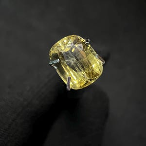 Puede incluir: Primer plano de un anillo con una piedra preciosa amarilla. El anillo presenta una gran piedra amarilla de corte rectangular engastada en una banda de metal plateado. La superficie facetada de la piedra preciosa refleja la luz, sobre un fondo oscuro.