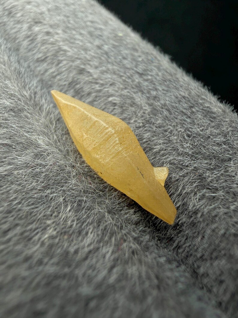 Puede incluir: Un peque&ntilde;o cristal amarillo claro con una punta y una superficie texturizada. El cristal est&aacute; sobre un fondo gris texturizado.