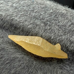 Puede incluir: Una peque&ntilde;a punta de flecha de piedra de color amarillo claro con una superficie texturizada. La punta de flecha tiene forma triangular con una peque&ntilde;a muesca en la base. El objeto est&aacute; sobre una superficie gris y peluda.