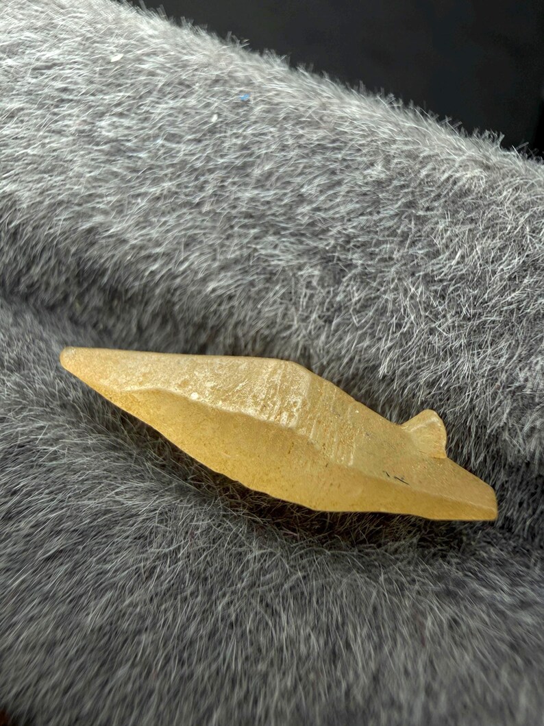 Puede incluir: Una peque&ntilde;a punta de flecha de piedra de color amarillo claro con una superficie texturizada. La punta de flecha tiene forma triangular con una peque&ntilde;a muesca en la base. El objeto est&aacute; sobre una superficie gris y peluda.