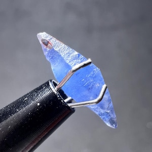 Puede incluir: Primer plano de una gema azul facetada sujeta por pinzas de metal plateado. La gema tiene una textura rugosa y natural y está sujeta por una herramienta negra sobre un fondo gris.