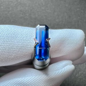May include: A deep blue gemstone set in a silver ring setting. The gemstone is a rectangular shape with a faceted cut.