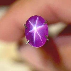 May include: A close-up of a purple star sapphire gemstone. The oval-shaped stone has a six-rayed star pattern in white, set in a silver metal setting. The gemstone is a vibrant shade of purple.