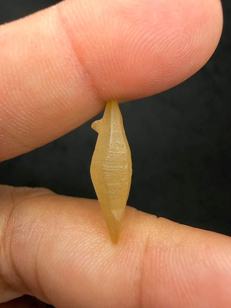 Puede incluir: Un peque&ntilde;o cristal transl&uacute;cido de color amarillo con una punta y una peque&ntilde;a protuberancia. El cristal se sostiene entre dos dedos sobre un fondo oscuro, mostrando su forma y textura.