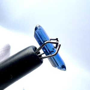 May include: A deep blue gemstone held in a metal clamp. The gemstone is a long, rectangular shape with pointed ends.