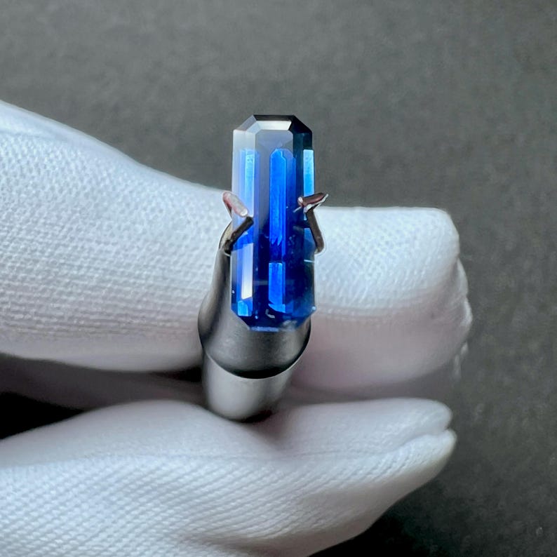 May include: A deep blue gemstone with a rectangular cut, held in a metal clamp. The gemstone has a slight shimmer and is a vibrant blue color.