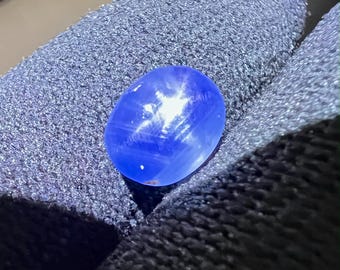 May include: A blue star sapphire gemstone with a star-like pattern in the center. The gemstone is oval-shaped and has a smooth, polished surface.
