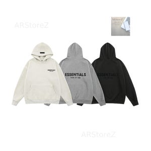 May include: Three hooded sweatshirts in off-white, grey, and black. Each sweatshirt has a front pocket and the word "ESSENTIALS" printed on the chest. The grey sweatshirt also has "FEAR OF GOD" printed below "ESSENTIALS".