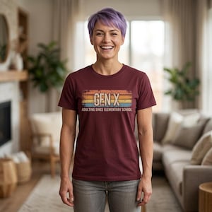Op de afbeelding: Een bordeauxrood t-shirt met de tekst "GEN X" in een retro-ontwerp, met de zin "ADULTING SINCE ELEMENTARY SCHOOL" eronder. Het t-shirt wordt gedragen door een persoon met kort paars haar en een grijze broek.