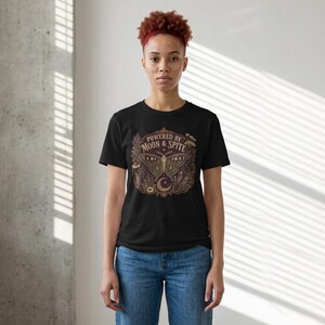 以下が含まれることがあります： 黒の半袖Tシャツ。蛾と植物のモチーフ、三日月が描かれたグラフィックプリントが施されています。蛾の上部には「POWERED BY MOON & SPITE」というテキストがアーチ状に明るい色でプリントされています。