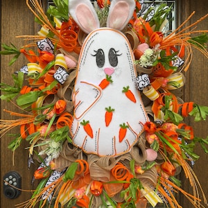 May include: A white bunny with pink ears and black eyes holding carrots is the centerpiece of this Easter wreath. The wreath is decorated with orange, yellow, and green ribbon, as well as green and orange tulle. The wreath is also decorated with small white flowers and green leaves.