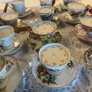 Puede incluir: Una colección de velas en tazas de té sobre platillos. Las tazas de té son blancas, florales y de colores pastel, con adornos dorados. Cada taza de té contiene una vela blanca con una sola mecha.