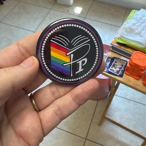 May include: A circular pin-back button featuring a black background with a white dotted border. The center displays an open book graphic with rainbow-colored pages and a stylized white letter P integrated into the design of the book pages.