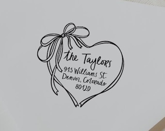 May include: A white envelope with a black heart-shaped design featuring a bow and the text "the Taylors" along with an address. The envelope is on a wooden surface.