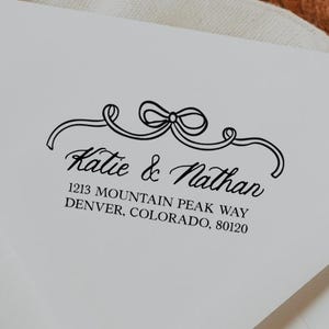 Op de afbeelding: Witte envelop met zwarte tekst. De tekst luidt "Katie & Nathan" en het adres "1213 Mountain Peak Way, Denver, Colorado, 80120". Boven de namen staat een decoratief strikdesign.