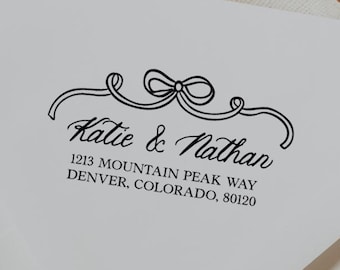 May include: White envelope with black text that reads "Katie & Nathan" and the address "1213 Mountain Peak Way, Denver, Colorado, 80120". The envelope has a decorative bow design above the names.
