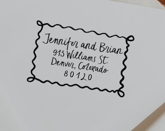 May include: A white envelope with a black, decorative border containing the text "Jennifer and Brian, 915 Williams St., Denver, Colorado 80120". The envelope is on a wooden surface.