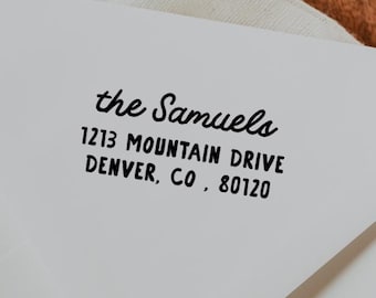May include: White envelope with the address "the Samuels, 1213 Mountain Drive, Denver, CO, 80120" printed in black. The envelope is on a wooden surface with a white cloth.