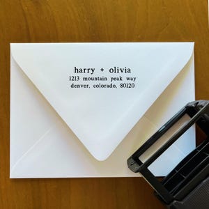 May include: A white envelope with the names "harry + olivia" and an address printed in black. The address includes the street name, city, state, and zip code. A black stamp is in the lower right corner.