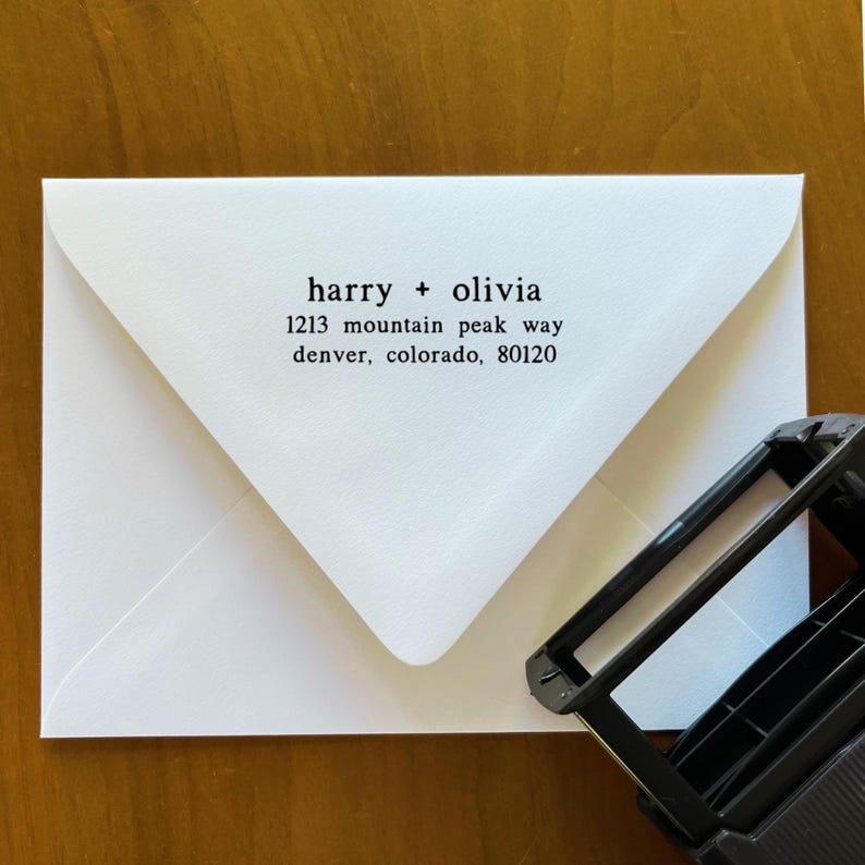 May include: A white envelope with the names "harry + olivia" and an address printed in black. The address includes the street name, city, state, and zip code. A black stamp is in the lower right corner.