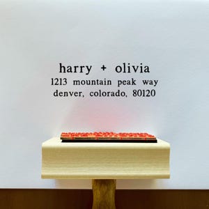 May include: A wooden address stamp with the names "harry + olivia" and address details in black ink. The stamp is resting on a white surface. The stamp's red ink pad is visible.