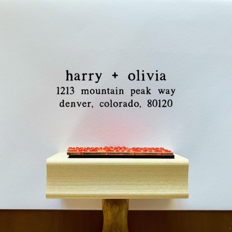 May include: A wooden address stamp with the names "harry + olivia" and address details in black ink. The stamp is resting on a white surface. The stamp's red ink pad is visible.