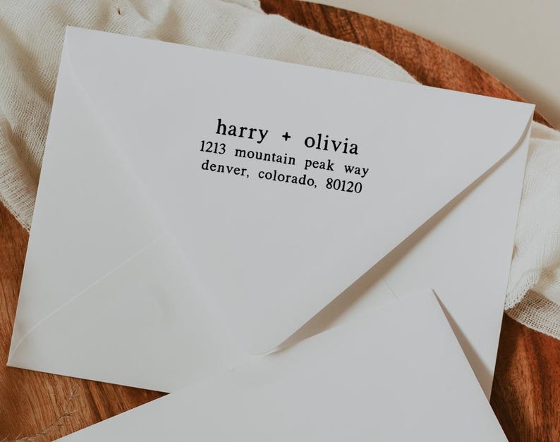 May include: White envelope with the text "harry + olivia" and address information in black. The envelope is on a wooden surface with a white cloth. The address reads "1213 mountain peak way, denver, colorado, 80120".