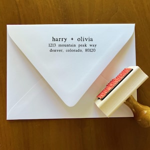 May include: A white envelope with the names "harry + olivia" and address details printed in black. A wooden stamp with a red ink pad is positioned next to the envelope, ready for use. The envelope is on a wooden surface.