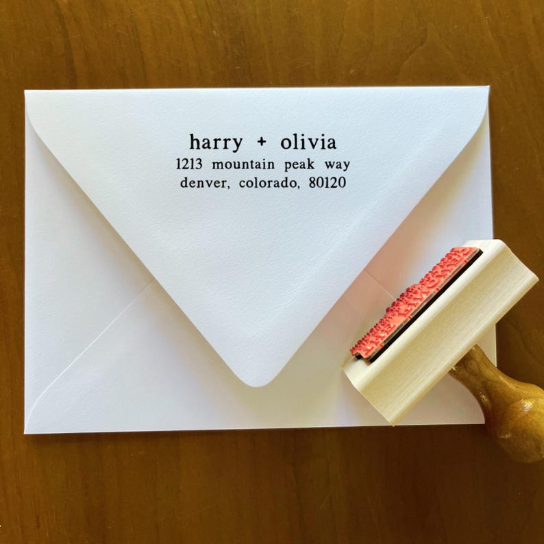 May include: A white envelope with the names "harry + olivia" and address details printed in black. A wooden stamp with a red ink pad is positioned next to the envelope, ready for use. The envelope is on a wooden surface.