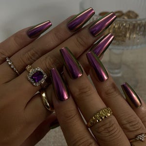 Op de afbeelding: Lange doodskistvormige press-on nagels met een metallic, iriserende paarse en groene chroomafwerking. De vingers zijn versierd met verschillende goudkleurige ringen, waaronder een met een grote rechthoekige paarse steen en heldere kristallen.
