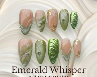 Peut inclure: Un ensemble de dix faux ongles aux designs et couleurs variés. Les ongles présentent un mélange de nuances nude, vertes et dorées, avec des détails de feuilles et de pierres précieuses. Le texte "Emerald Whisper" et "Autumn Collection" est affiché.