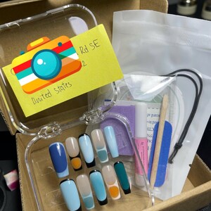以下が含まれることがあります： 黒のチップとオレンジとターコイズのアクセントが付いた、ブルー、ターコイズ、ベージュの付け爪のセットです。爪は、「Rd SE 2 United States」と書かれた黄色の付箋が付いた透明なプラスチックケースに梱包されています。パッケージには、小さな接着剤のボトル、爪やすり、木製のキューティクルプッシャー、小さなネイルステッカーの袋も含まれています。
