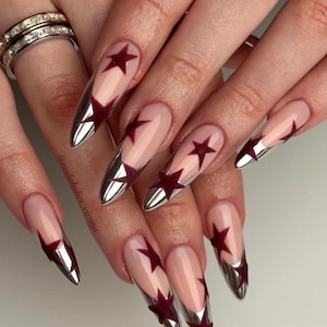 May include: Long almond-shaped nails featuring a nude base, metallic silver chrome tips, and dark red star-shaped accents. The design includes a clear coat finish, and the text @nailaholics_vienna is visible on the side of one fingernail.
