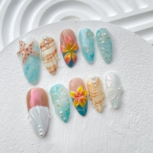 Könnte beinhalten: Zehn verschiedene Nageldesigns mit Muscheln, Seesternen und Blumen. Die Nägel sind in Blautönen, Weiß und Rosa lackiert.