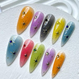 Puede incluir: Diez u&ntilde;as postizas largas en forma de almendra con efecto degradado. Cada u&ntilde;a est&aacute; pintada en un color diferente, incluyendo azul, verde, amarillo, naranja, rojo, morado y gris. Cada u&ntilde;a tiene un dise&ntilde;o de estrella dorada y un peque&ntilde;o strass.